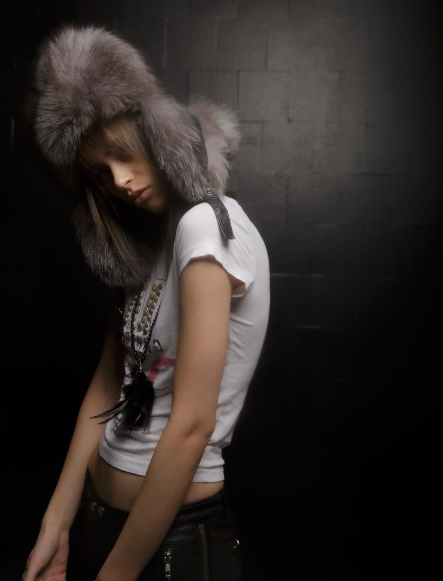 Editorial fashion photography — model in an oversized fur hat against a dark brick wall, low-light portrait shoot by Maison Division